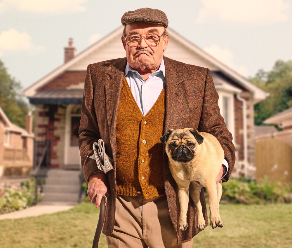 Elderly man in tweed suit holds a pug outdoors near a house. He holds a cane and newspaper. Sunny, eye-level medium shot.