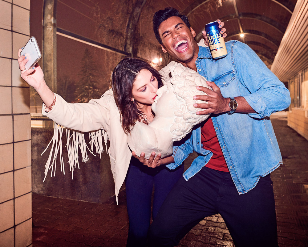 Woman kisses sculpture as man holds beer in tunnel, taking selfie with smartphone. Beige tile, concrete, and beverage branding visible.