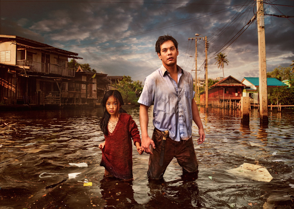 Flooded street scene featuring an adult male and a child, standing in waist-deep water with houses and powerlines in the background.