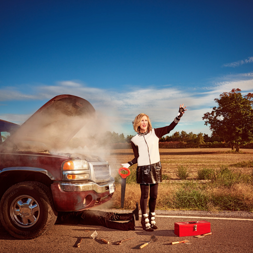 Woman in fashion attire stands by a smoking pickup truck on a country road, holding a red toolbox, against a rural background and blue sky.