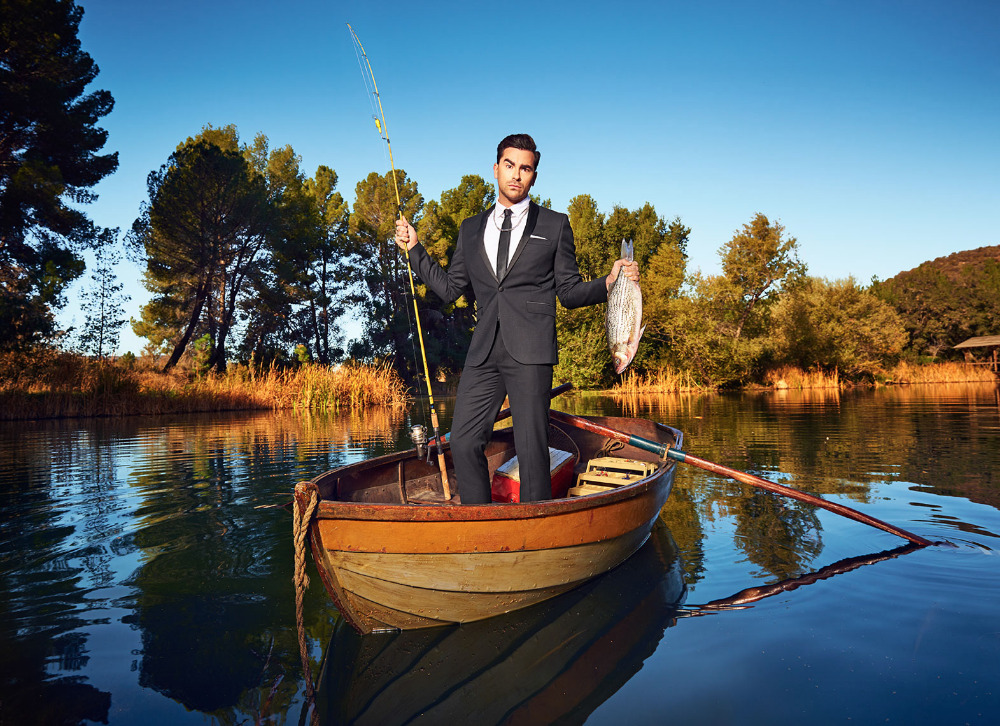 Man in suit on wooden rowboat holds a fish and fishing pole on lake surrounded by trees under a clear blue sky.