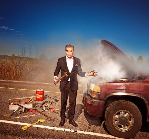 Adult male in suit stands near smoking car on roadside holding wrench, surrounded by tools under clear sky. Vehicle trouble concept.