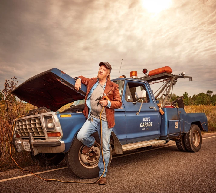 An adult male leans against a blue tow truck with its hood open, holding jumper cables, in a rural setting under a cloudy sky.
