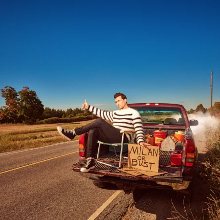 Adult male hitchhiking from the back of a red pickup truck on a rural road under a bright blue sky with a cardboard sign.