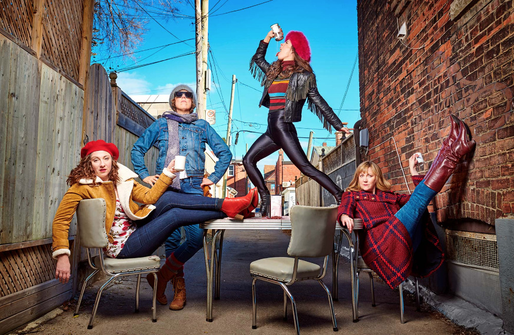 Four women strike unique poses at an outdoor cafe, each holding a cup. They showcase vintage fashion in an urban alley setting.