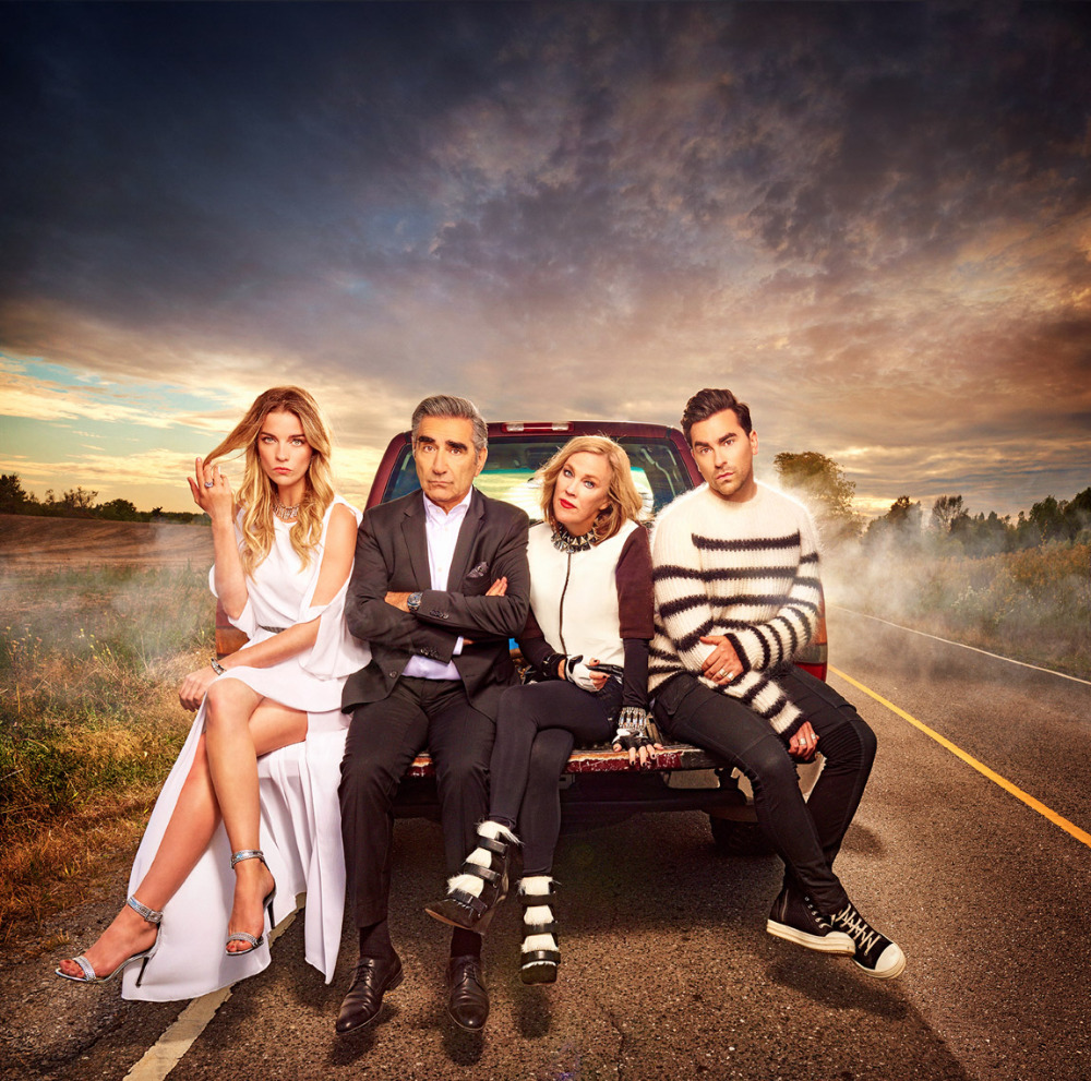 Four adults sit on the back of a pickup truck on an asphalt road with an open field under a dramatic, cloudy sky at dusk.