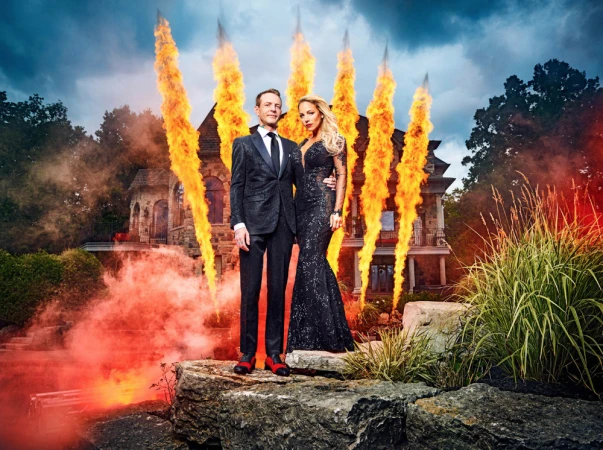Glamorous couple poses on rocks before a large house with special effects of fire, wearing formal attire at dusk.