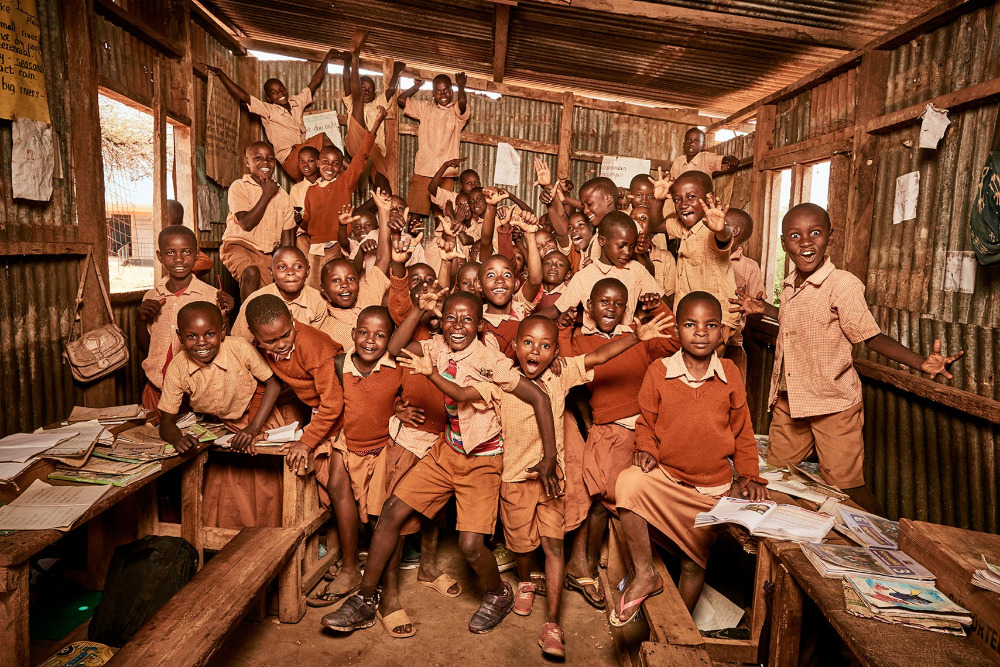 Group of happy African children in school uniforms pose together inside a corrugated metal classroom, desks covered with books.