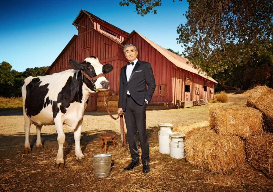 Man in tuxedo with cow in rural setting, standing in front of red barn with bales of hay and metal buckets. Clear sky.