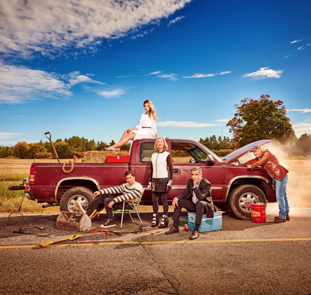 Group of five people gathered around a maroon pickup truck on a rural road under a blue sky with hay bales and trees in the background.