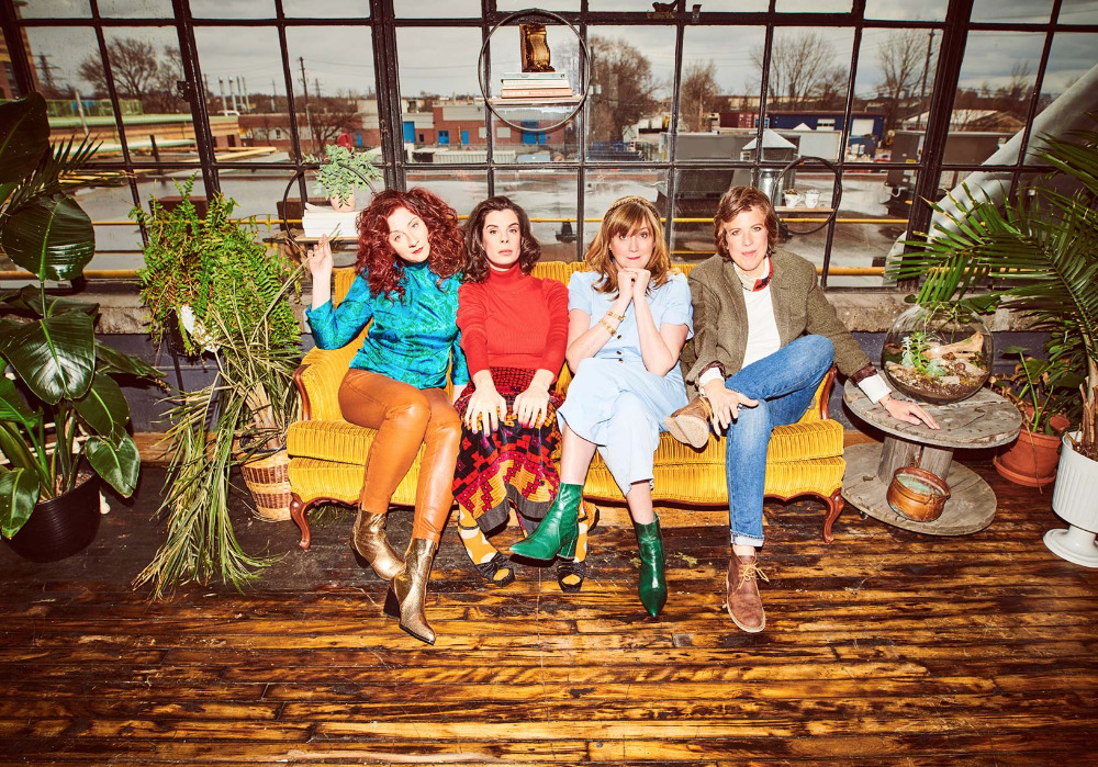 Four women pose on a yellow couch indoors, surrounded by plants. The background features a window and urban scenery with industrial buildings.