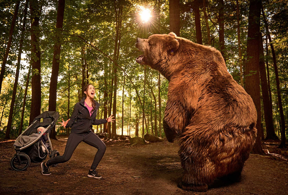 Woman with stroller reacts to a bear in the woods. The forest setting has tall trees and bright sun, creating a suspenseful scene.