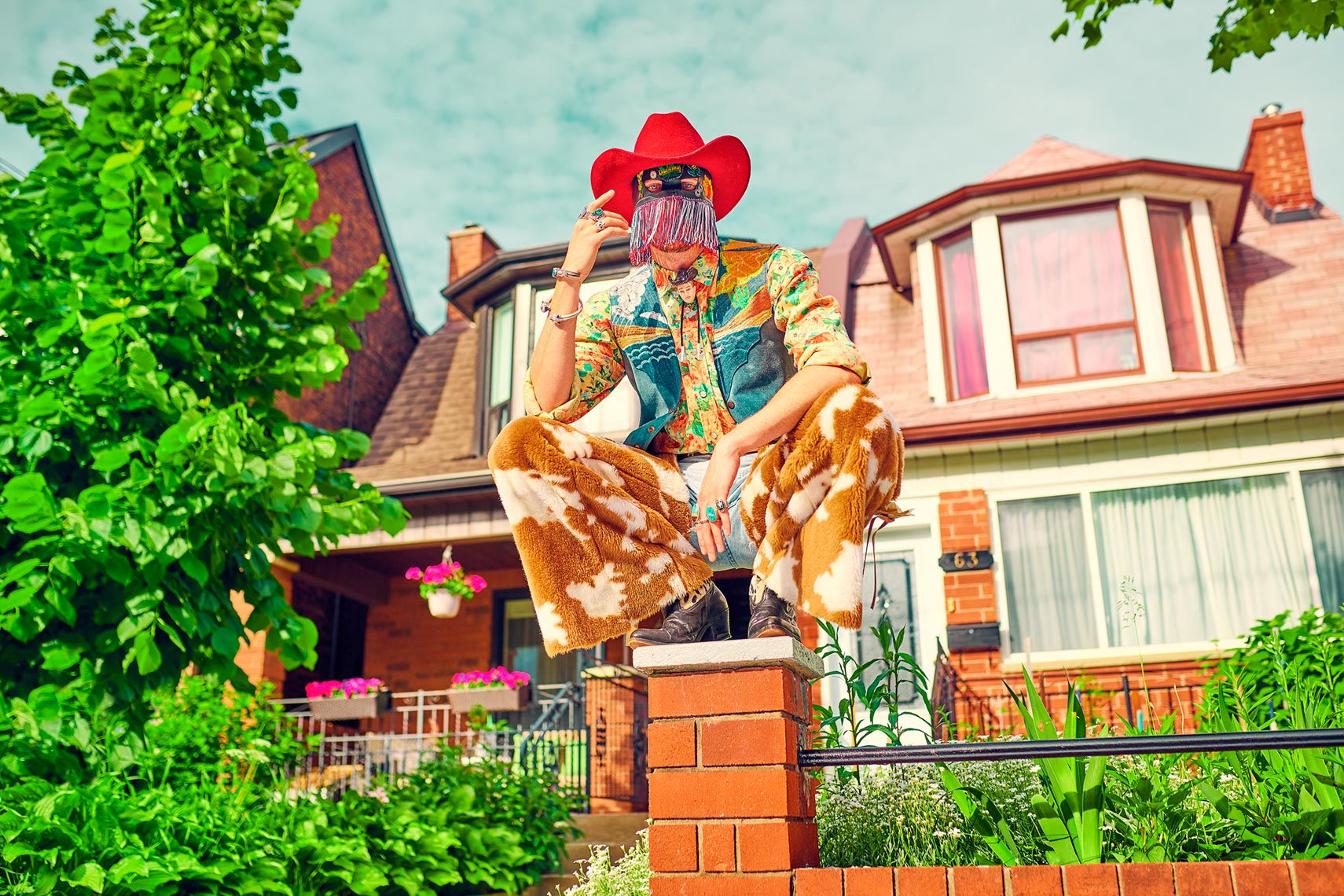 Person in cowboy hat and patterned outfit crouching on a brick fence in front of a house with trees and blue sky.