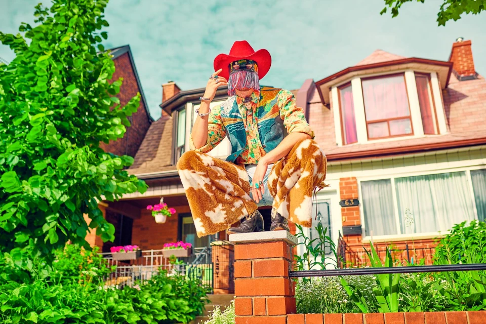 Person in cowboy hat and patterned outfit crouching on a brick fence in front of a house with trees and blue sky.