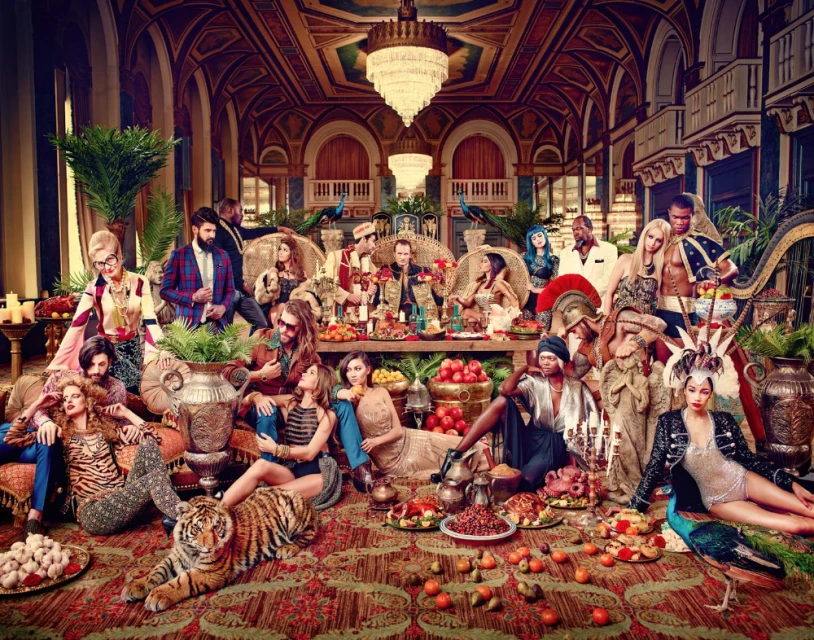 A diverse group of men and women in formal and ornate attire gather for a feast in a lavish, classically styled interior setting.