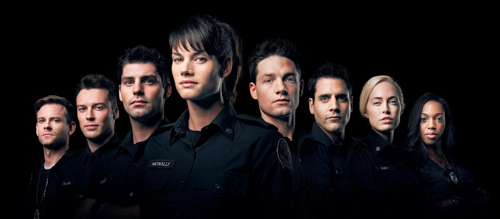 Group portrait of eight adult men and women wearing dark shirts, posed closely together against a solid black background.