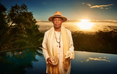 Elderly black woman wearing a tan fedora hat and cream tunic holding oyster mushrooms in front of an outdoor pool at sunset