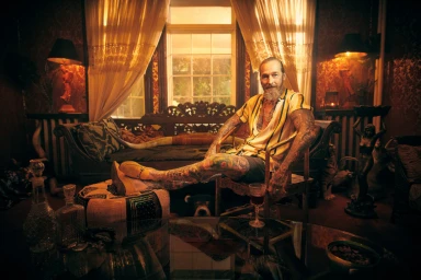 An elderly bearded man with extensive tattoos sitting in a lavishly decorated room, captured by Matt Barnes