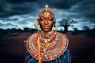 Portrait of a Black woman wearing elaborate beaded necklaces and a silver headdress under a dramatic stormy sky.
