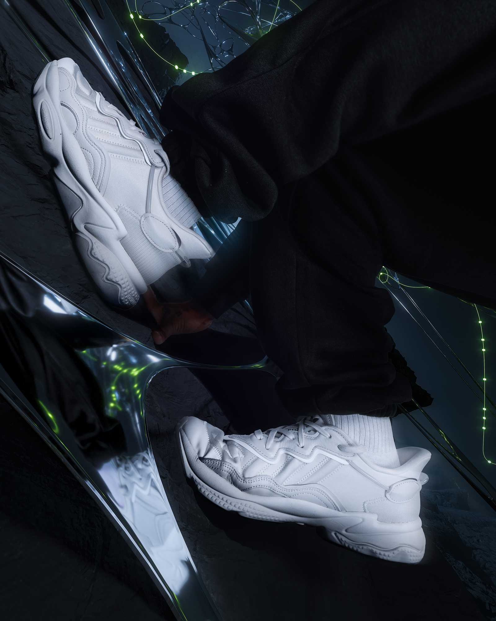 Two white Adidas Ozweego sneakers are showcased on a reflective silver surface with black fabric, creating a futuristic vibe.