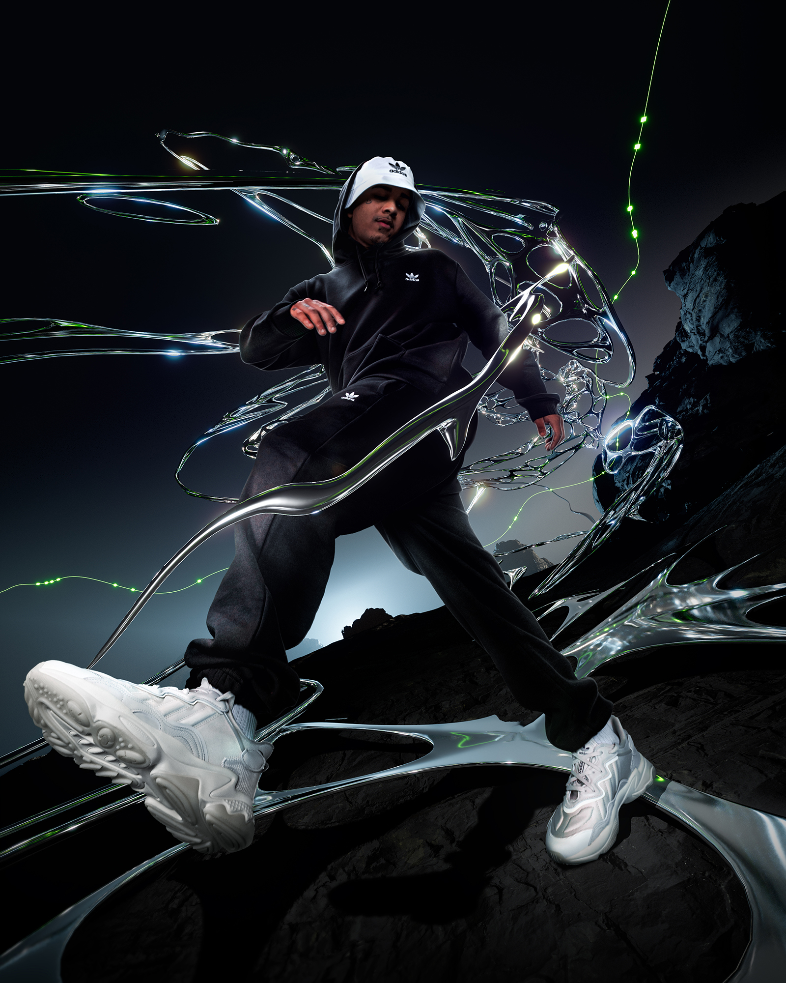 A person poses in a black Adidas tracksuit and white sneakers on a black, reflective surface with metallic accents in a studio setting.