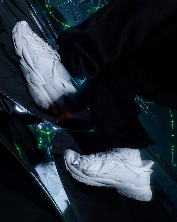Close-up of white chunky sneakers and black pants positioned on a reflective chrome surface in a dark futuristic environment.
