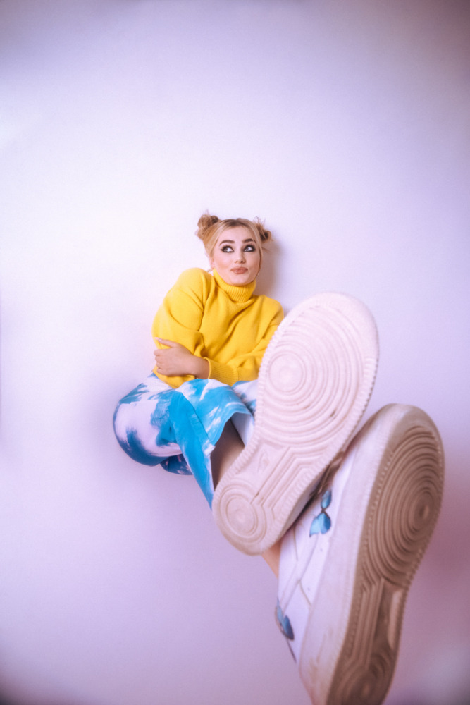 Blond woman in yellow sweater and tie-dye pants poses with white sneakers against a plain white backdrop in a vertical portrait.