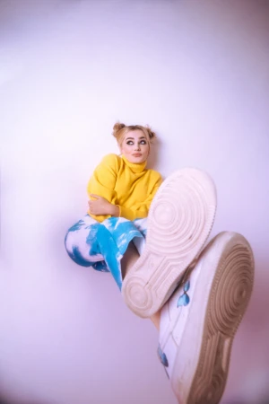 Blond woman in yellow sweater and tie-dye pants poses with white sneakers against a plain white backdrop in a vertical portrait.