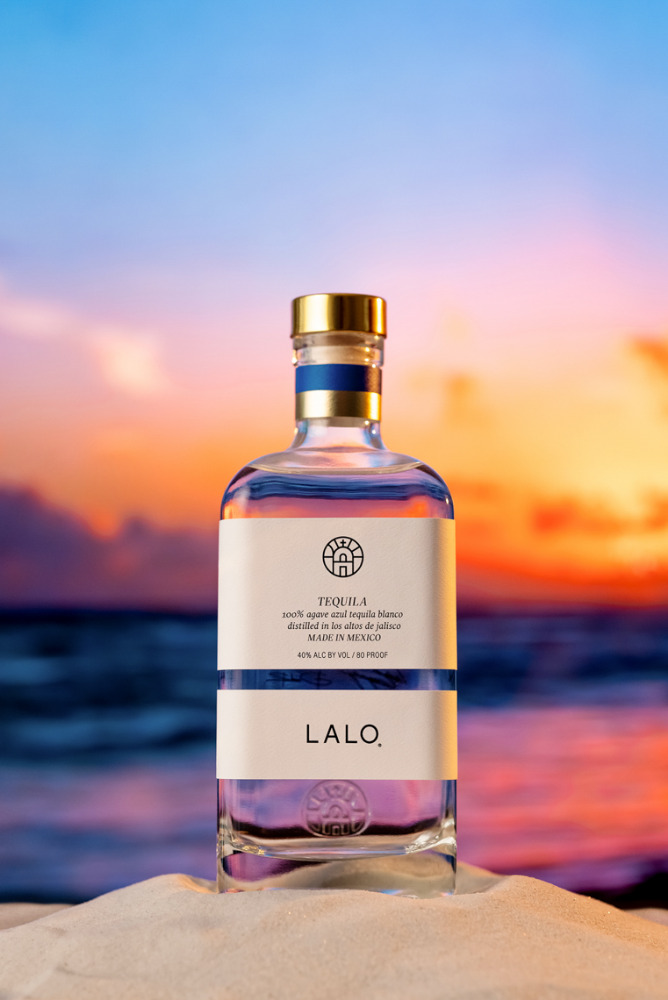 Lalo tequila bottle stands on a sand dune with a colorful sunset and ocean in the background creating a tropical, beachy vibe.