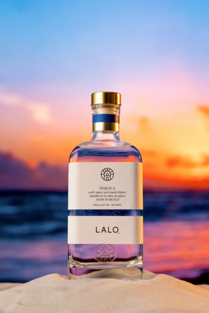 Lalo tequila bottle stands on a sand dune with a colorful sunset and ocean in the background creating a tropical, beachy vibe.