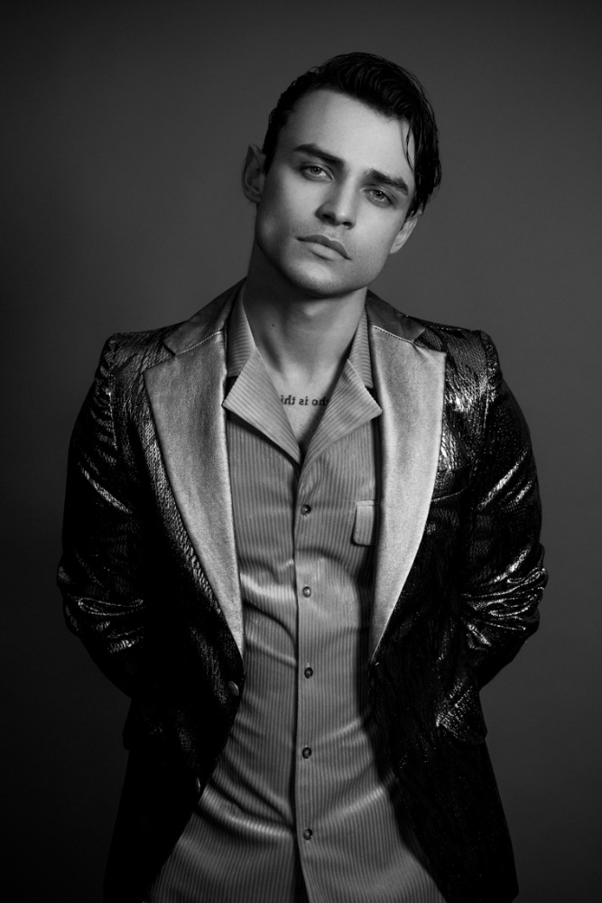 Monochrome portrait of a young adult male wearing a shiny blazer and striped shirt, set against a plain grey studio backdrop.