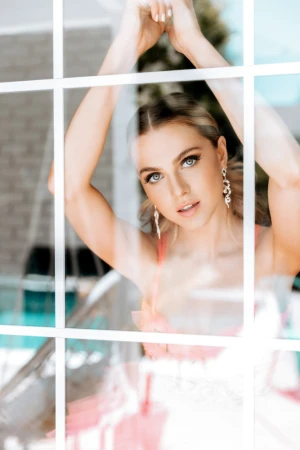 Woman posing behind a white window frame with arms raised, wearing drop earrings and a pink top.
