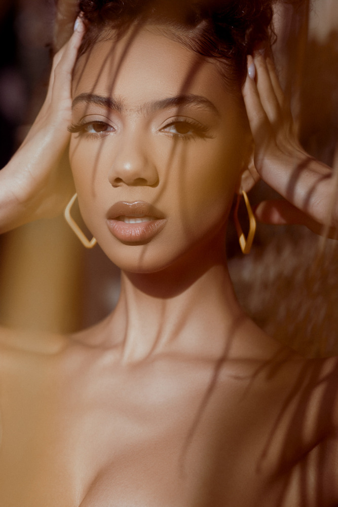 Close-up portrait of a biracial young adult woman with hands framing face, gold earrings, and shadows, capturing beauty and elegance.
