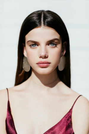 Portrait of a woman with dark hair and blue eyes wearing large disc earrings against a white background.