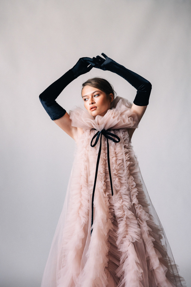 Woman in a pale pink ruffled dress with black velvet gloves poses elegantly in a studio with arms raised above her head.