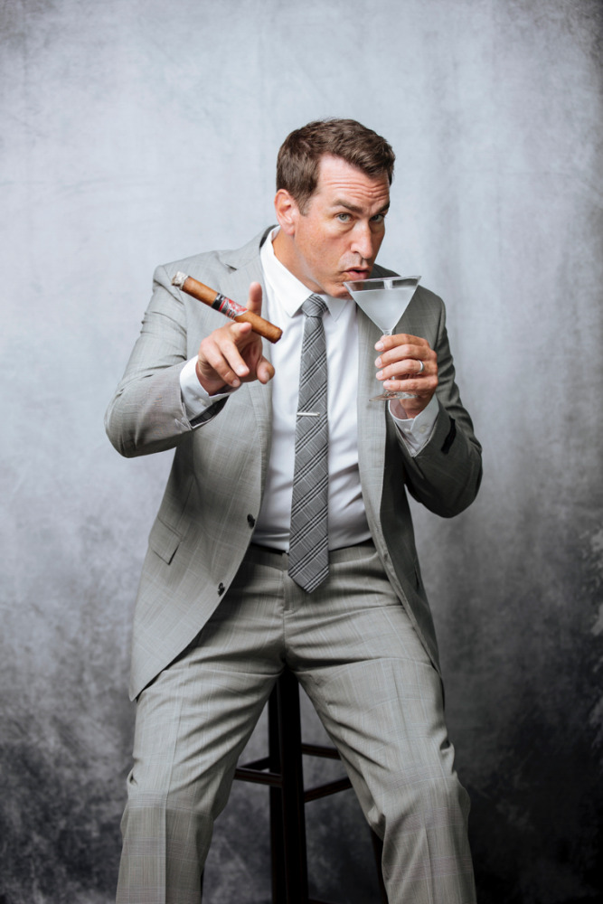 Man in suit sits holding a cigar and martini glass against a grey backdrop in a studio shot. Light grey suit and tie.