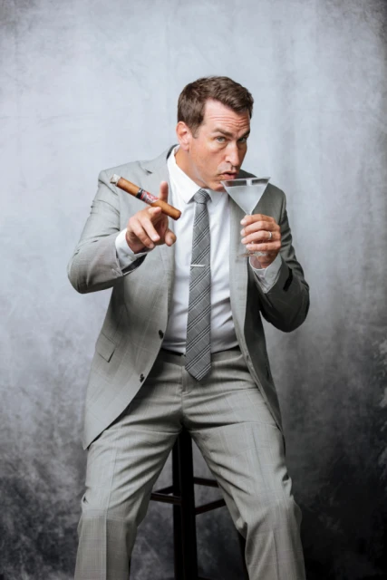 Man in suit sits holding a cigar and martini glass against a grey backdrop in a studio shot. Light grey suit and tie.