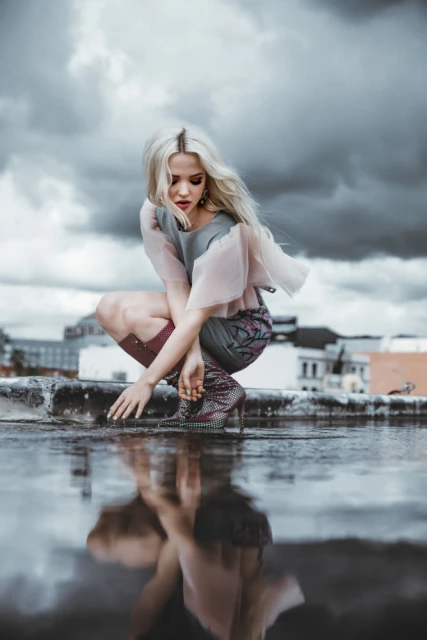 A young woman with blonde hair squats on a rooftop next to a puddle reflecting her image, with a cityscape and cloudy sky in the background.