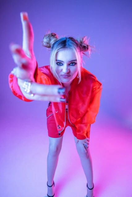 Blonde woman in red jacket poses in a photo studio with dynamic lighting, showing playful attitude and stylish makeup, set against a vivid backdrop.