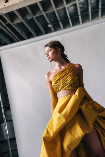Woman in a mustard yellow gown with a bare shoulder poses in a photo studio in front of a white backdrop and under industrial ceiling.