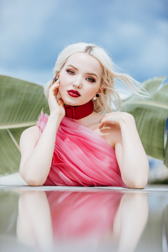 Close-up portrait of a blonde adult female with red lipstick, wearing a pink dress and red collar, posing outdoors with green foliage.
