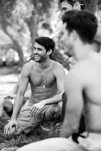 Black and white portrait of a happy shirtless man sitting outdoors with a group of friends in a blurred nature setting.