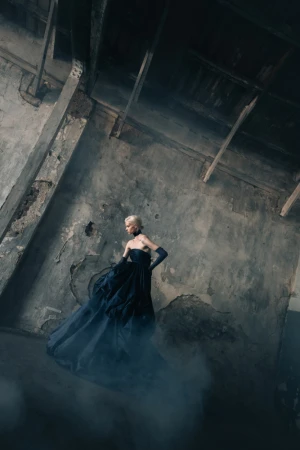A blonde model wearing a dark strapless gown and long gloves poses in a weathered industrial interior.