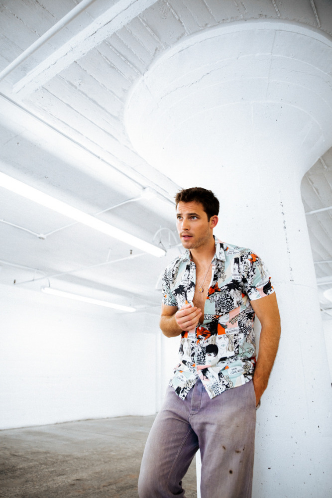 Portrait of an adult male leaning against a pillar wearing a patterned short-sleeve shirt and grey pants in a bright indoor space.