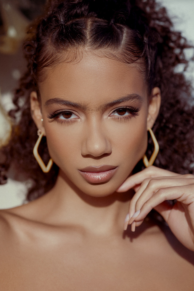 Close-up portrait of a young woman with curly hair, wearing geometric earrings, posing indoors in soft, diffused lighting for a beauty shot.