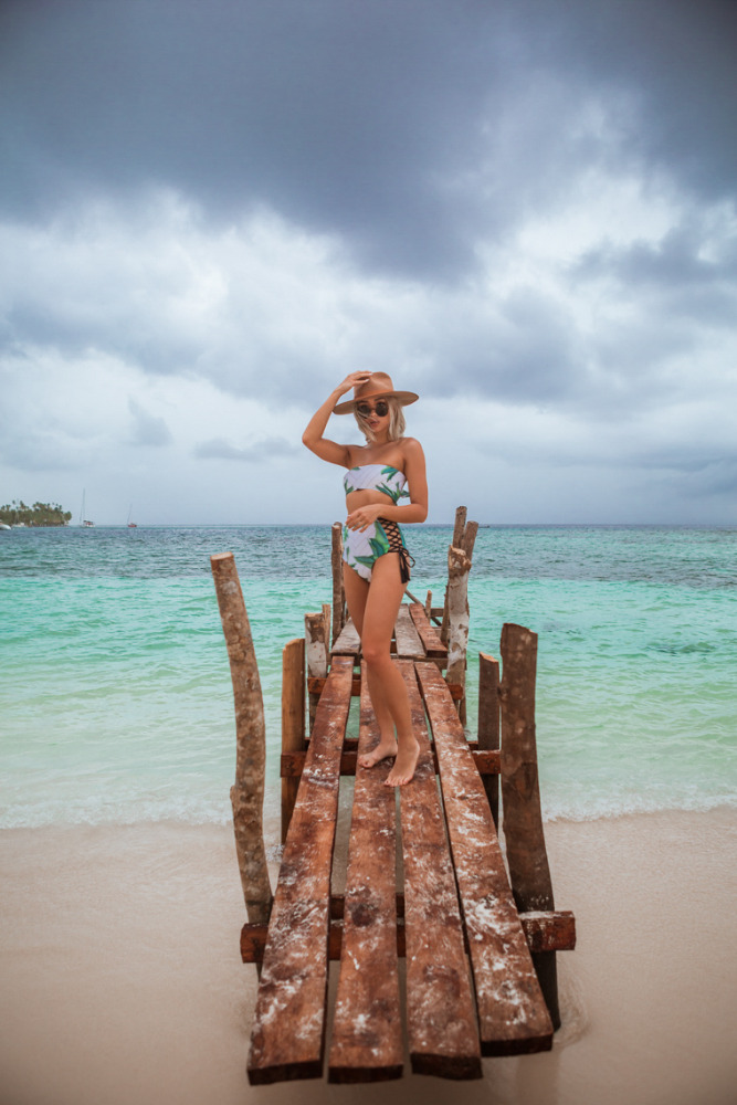 A woman in a bathing suit and sun hat stands on a rustic wooden dock over clear, blue water on a cloudy day at a tropical beach.