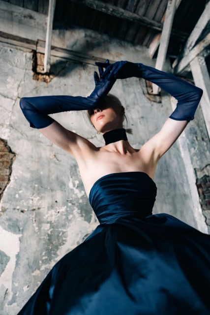 A low-angle portrait of a fair-skinned young woman in a navy blue dress and opera gloves, posing in a distressed, abandoned space.