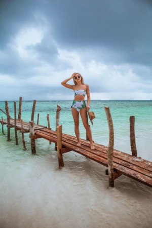 Woman in floral bikini on wooden pier at tropical beach destination with cloudy sky, holding sunglasses and hat. Travel fashion concept.
