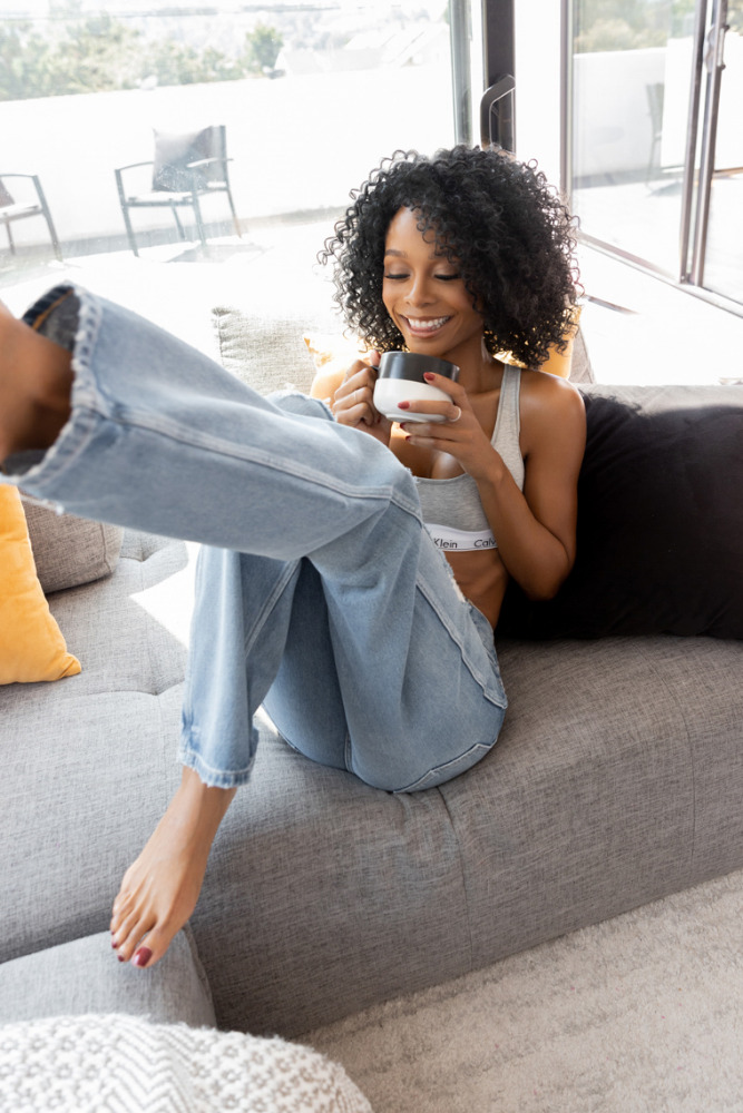 A woman with curly hair wearing a Calvin Klein bra and jeans relaxes on a gray sofa while holding a coffee mug in a sunlit interior.