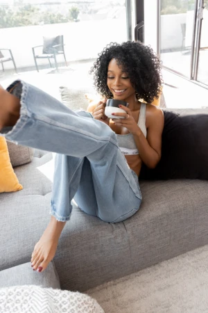 A woman with curly hair wearing a Calvin Klein bra and jeans relaxes on a gray sofa while holding a coffee mug in a sunlit interior.